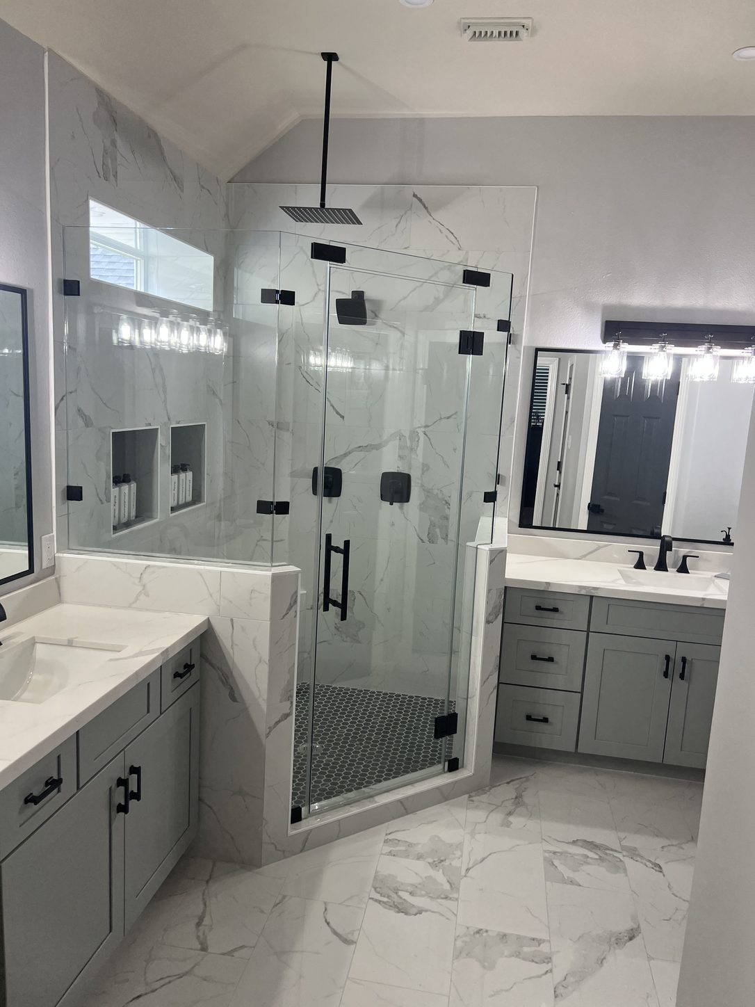 The remodeled bathroom from the doorway: a glass-enclosed walk-in shower between two vanities with gray cabinetry and matte black hardware, on veined white tile